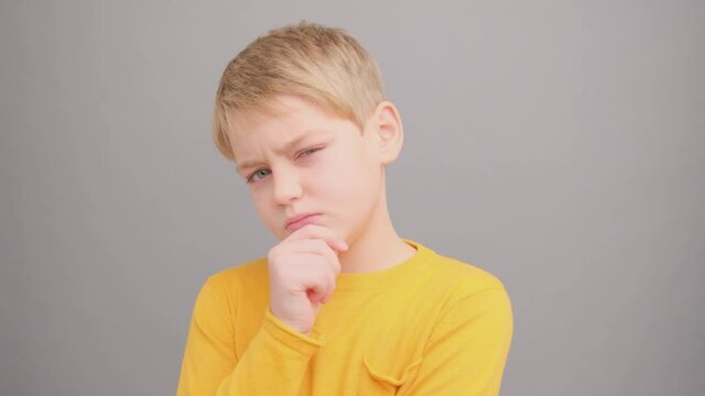 Little boy is thoughtful and looks at the camera in the studio on a gray background. Super slow motion.