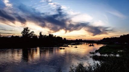 Fototapeta na wymiar incredible mysterious amazing swirls of clouds at sunset on the river with reflection in the water on a warm summer evening. in the background - a bridge over the river and a tent camp on the river