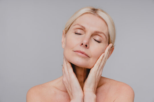 Beautiful aged white woman touching her clean pure face with her eyes closed isolated over grey background