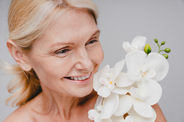 Close up portrait of middle-aged beautiful woman with orchid branch isolated over grey background