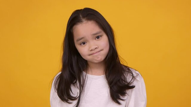 Cute eastern girl shrugging shoulders and spreading hands, feeling unsure and doubtful, orange studio background