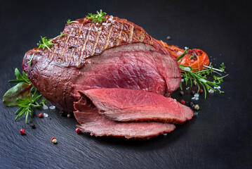 Modern style traditional barbecue haunch of venison sliced and as piece served with tomatoes and herbs as close-up on a black board - obrazy, fototapety, plakaty