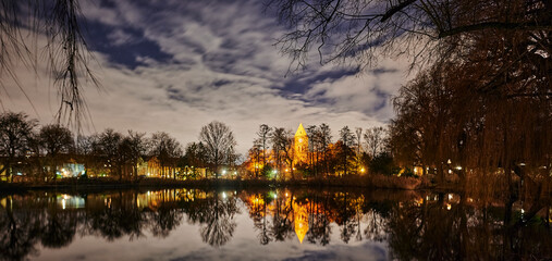 A historic village church that is illuminated by lamps in Berlin-Lichtenrade at night and that is reflected in a pond.