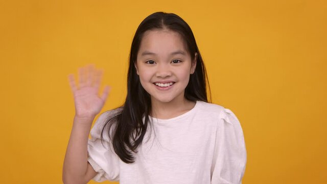 Cute friendly asian girl waving hand hi and smiling to camera, orange studio background