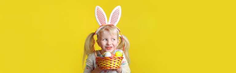 Caucasian blonde girl with pink Easter bunny ears holding eggs. Person in studio on a yellow background. Funny kid child celebrating traditional Christian holiday. Web banner header.