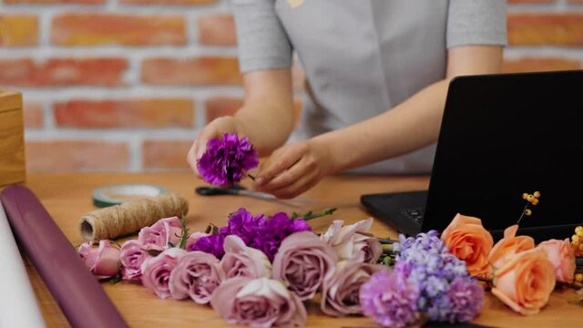Floral store. Close up hands woman florist. Female florist in apron at workplace touch flowers and typing in notebook. Delivery online. Small business owner working on laptop in store. Slow motion.