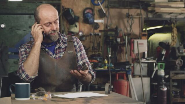 Male blacksmith working on design in forge whilst talking on mobile phone - shot in slow motion