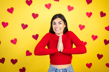 Young caucasian woman over yellow background with red hearts begging and praying with hands together with hope expression on face very emotional and worried
