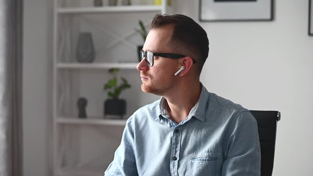 Thoughtful young businessman wearing modern stylish glasses and smart casual shirt looks away and worried about some task, solving problem. A pensive male employee with a airpods in ears