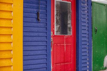 Fototapeta na wymiar A bright red metal door with a clear glass closed window. The entrance has a deep royal blue wood clapboard wall with a bright yellow edging. There's a green wooden shed door with a black metal latch.