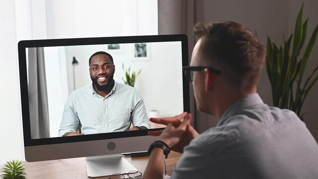 Video conference concept. Two colleagues caucasian and african men is talking via video connect, an African-American guy smiles on the laptop screen, chatting online. View above shoulder