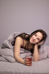 charming sleepy brunette woman with glass cup of tea in bed with grey bed linens. morning routine.
