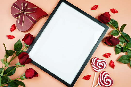Happy Valentine's Day black border picture frame, styled with Valentine red roses, heart shape gift and lipstick kissess chocolates on a modern coral background. Mockup. Top view flat lay. Copy space.