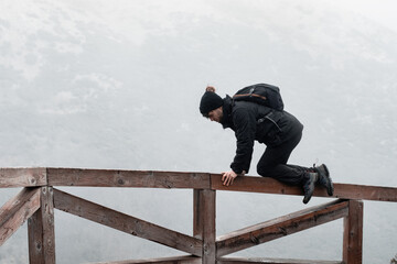 The young hiker dressed in black winter clothes climbs a wooden fence next to a mountain.