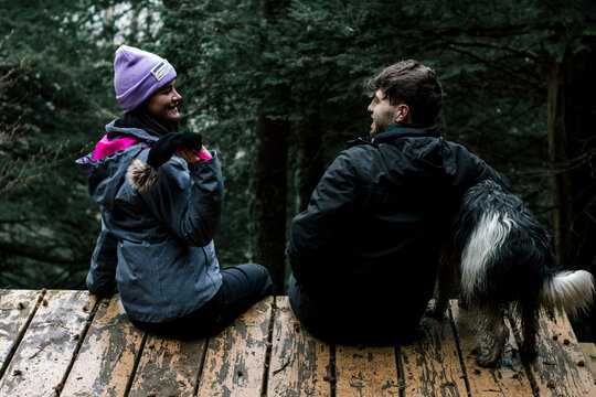 The Young Couple Are Sitting With Their Backs To The Camera On A Wooden Plank Road In The Forest. The Young Woman Takes Off Her Boyfriend's Hat.