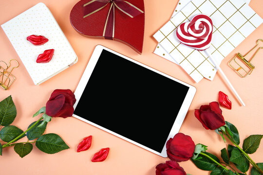 Happy Valentine's Day desktop with tablet device, styled with Valentine red roses, heart shape gift and lipstick kissess chocolates on a modern coral background. Mockup. Top view flat lay. Copy space.