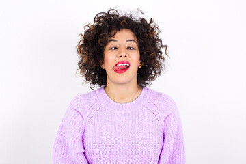 Funny young beautiful caucasian woman wearing pink knitted sweater against white wall makes grimace and crosses eyes plays fool has fun alone sticks out tongue.