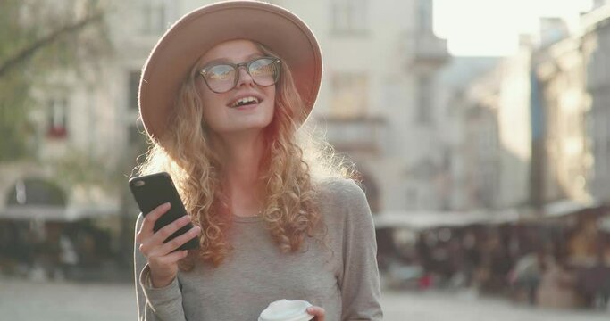 Portrait of Smiling and Satisfied Woman having a Walk in the Old Part of City. Reading funny Messages on her Smartphone. Holding paper cup with Coffee to go. Having trendy Hat and Charming Appearance.
