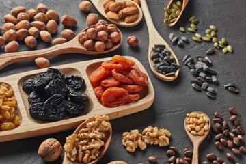 Different kinds of nuts, dried fruits in wooden spoones and dish on black slate background. Top view. Healthy food. Vegetarian nutrition