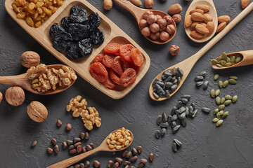 Different kinds of nuts, dried fruits in wooden spoones and dish on black slate background. Top...