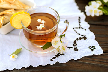 Glass cup with tea and petals, lemon, croissants, embroidered napkin, jasmine branches on a wooden table. Outdoor, picnic, brunch. Selective focus with short DOF. Close up view.
