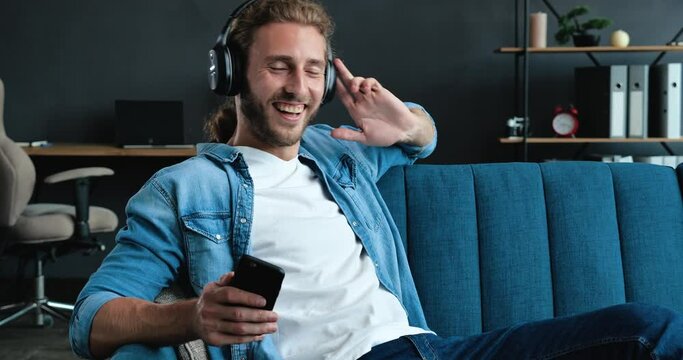 Caucasian Bearded Hipster on the bed and listening to Music in Headphones. Young Man enjoying cool Music on Couch in Loft Living Room. Playfully Mood at Home. FunnyMan. Hobby. Apps.