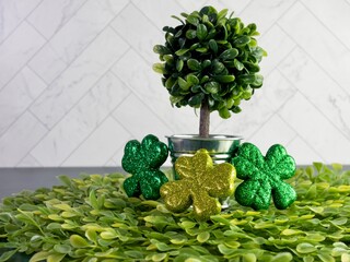 Potted tree sitting on a bed of green leaves with 3 four leaf clovers covered in glitter, against a...