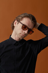 Attractive elegant young man with long hair in sunglasses, wear black shirt, standing on brown studio background.