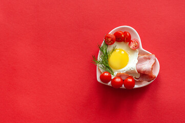Valentine's Day. A plate in the shape of a heart with scrambled eggs, bacon and tomatoes on a red background. Festive breakfast.