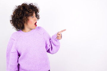 Stunned young beautiful caucasian woman wearing pink knitted sweater against white wall with greatly surprised expression points away on copy space, indicates something