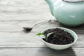 Dry black tea leaves in bowl and teapot on wooden background