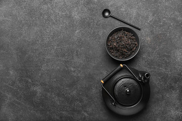 Obraz na płótnie Canvas Dry black tea leaves in bowl, teapot and spoon on dark background