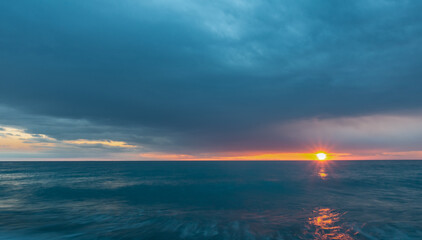 Fototapeta premium Dense blue clouds and turquoise sea. Between the sky and the water, the setting sun illuminates the sky orange. Sunny path. Black Sea