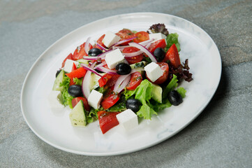 Close up of Traditional Greek salad on marble plate. Salad with Feta cheese, black olives, lettuce, fresh cucumber, cherry tomatoes served with onion on stone background
