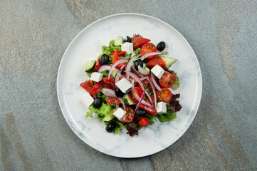 Top view of Traditional Greek salad on marble plate. Salad with Feta cheese, black olives, lettuce, fresh cucumber, cherry tomatoes served with onion on stone background. Minimalistic food serving

