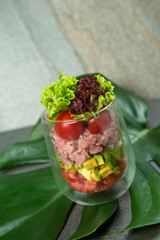Raw tuna fish tartare salad served in transparent glass. Layers of vegetables: tomato, avocado cubes, lettuce and cherry tomato. Seafood starter dish on stone background with tropical monstera leaf
