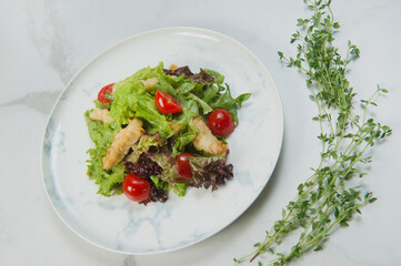 Side view of Chicken Tempura Salad served on white plate. Marble background with fresh thyme herbs. Meat Dish with deep fried chicken strips, lettuce and cherry tomatoes
