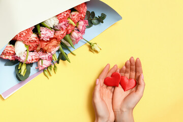 A bouquet of roses and female hands are holding two hearts in the palms on a yellow background. Valentine's day concept, symbol of love, romance.