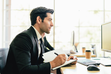 The Caucasian businessman jot something on small notebook while looking on computer monitor at his desk workplace.