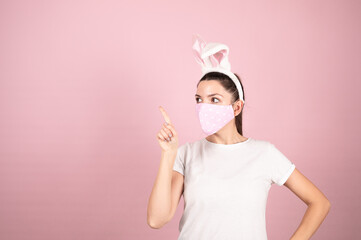 Young woman wearing bunny ears and protective mask standing over isolated pink background cheerful presenting and pointing with palm at something
