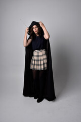 full length portrait of pretty brunette woman wearing tartan skirt and boots with long black cloak.. Standing pose holding a magical wand against a  studio background.