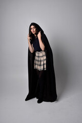 Naklejka premium full length portrait of pretty brunette woman wearing tartan skirt and boots with long black cloak.. Standing pose holding a magical wand against a studio background.