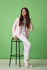 brunette teen girl in a pink jacket and white jeans posing sitting on a high stool on a green background