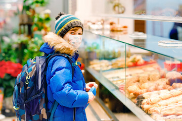 Kid boy wearing medical mask buy bread and pastry for school lunch in bakery. Child with backpack...