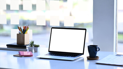Mock up laptop computer with white screen, coffee cup and office supplies on white table.