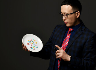 Young asian businessman pharmacist doctor in checkered official suit, tie and glasses holds shows plate of medical pills explaining treatment over dark background. Drugs, medicare, healthcare concept