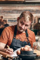 Caucasian man in a apron writes the recipe into the diary. Life style photography. Home kitchen. Vertical shot