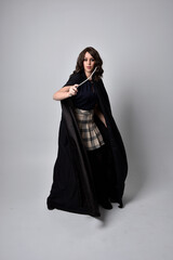 Naklejka premium full length portrait of pretty brunette woman wearing tartan skirt and boots with long black cloak.. Standing pose holding a magical wand against a studio background.