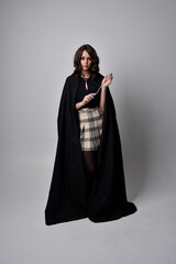 Fototapeta premium full length portrait of pretty brunette woman wearing tartan skirt and boots with long black cloak.. Standing pose holding a magical wand against a studio background.