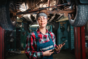 Fototapeta na wymiar A young beautiful female mechanic, in a uniform and glasses, with a tablet in her hands, poses standing under a car on a lift